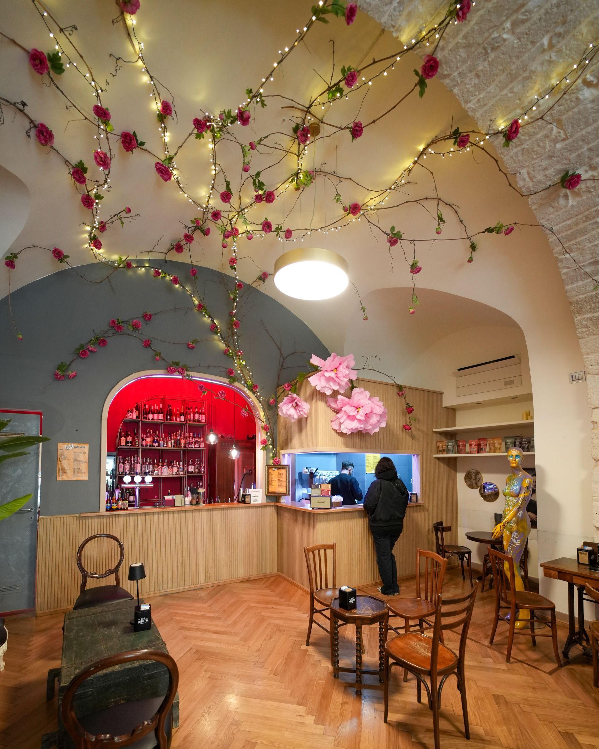 Interno del bar con tralci di fiori rosa, soffitto a volta, bancone e pavimento in legno. Persona al bancone.