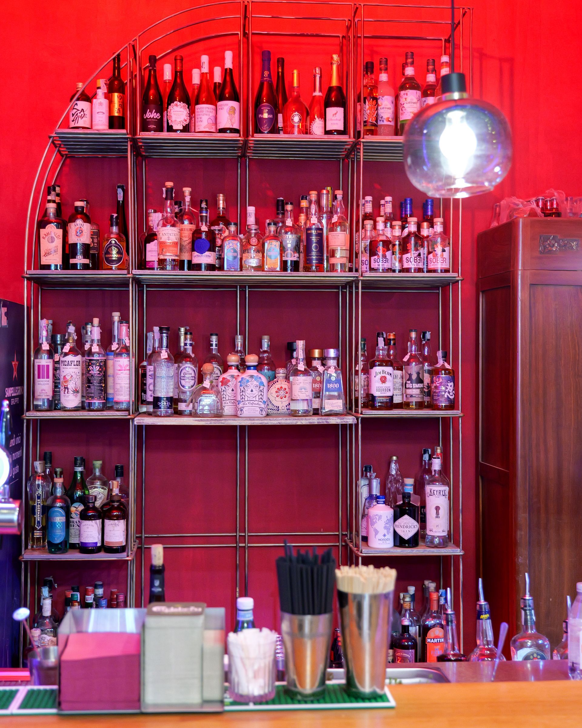 Scaffali pieni di bottiglie di liquore in un ambiente da bar, sfondo rosso, ripiani in acciaio inossidabile e una grande lampada rotonda.