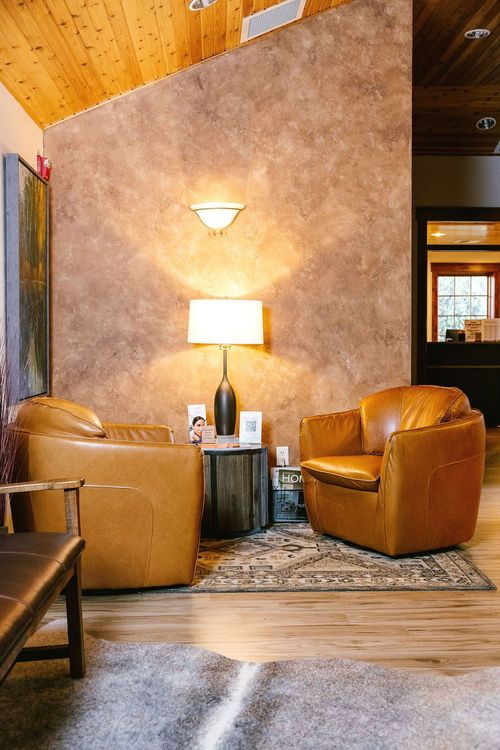 Two leather chairs in a waiting area with textured brown wall, table, lamp, and rug.