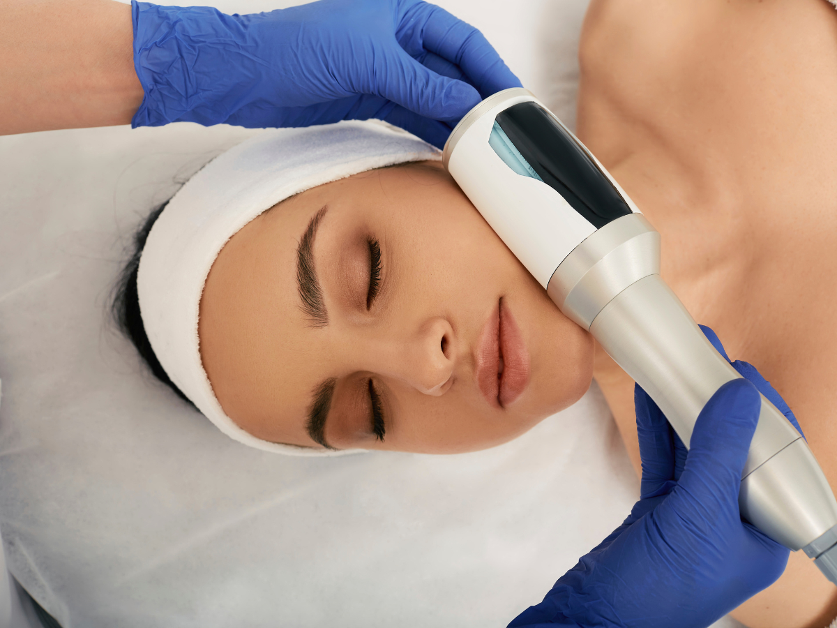 Woman receiving facial treatment; device held to cheek by gloved hands.