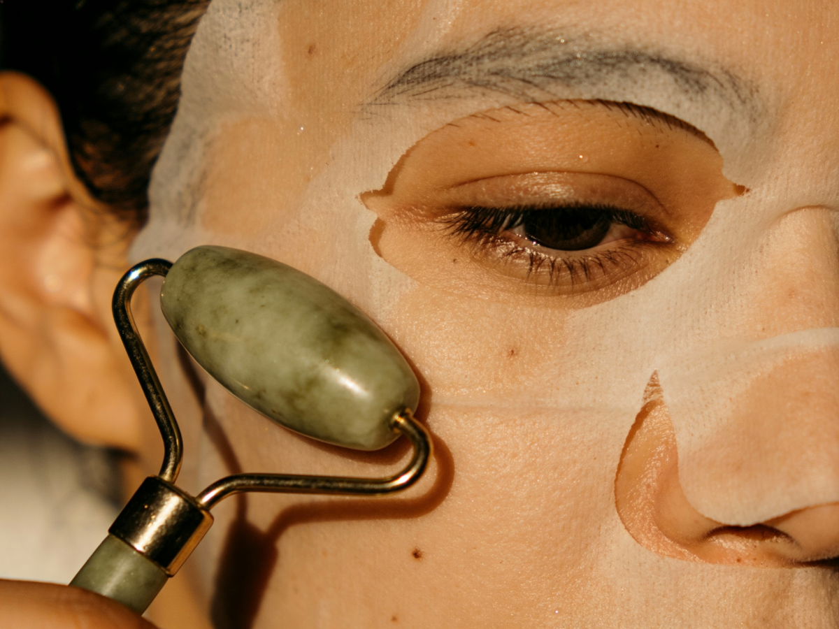 Person applying jade roller to face, wearing a sheet mask.