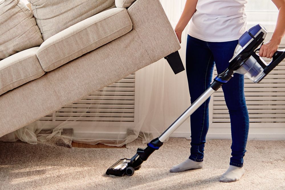 Person Vacuuming Carpet Near a Lifted Sofa in a Room With a Window — Clean Coast Carpet Cleaning in Port Stephens, NSW