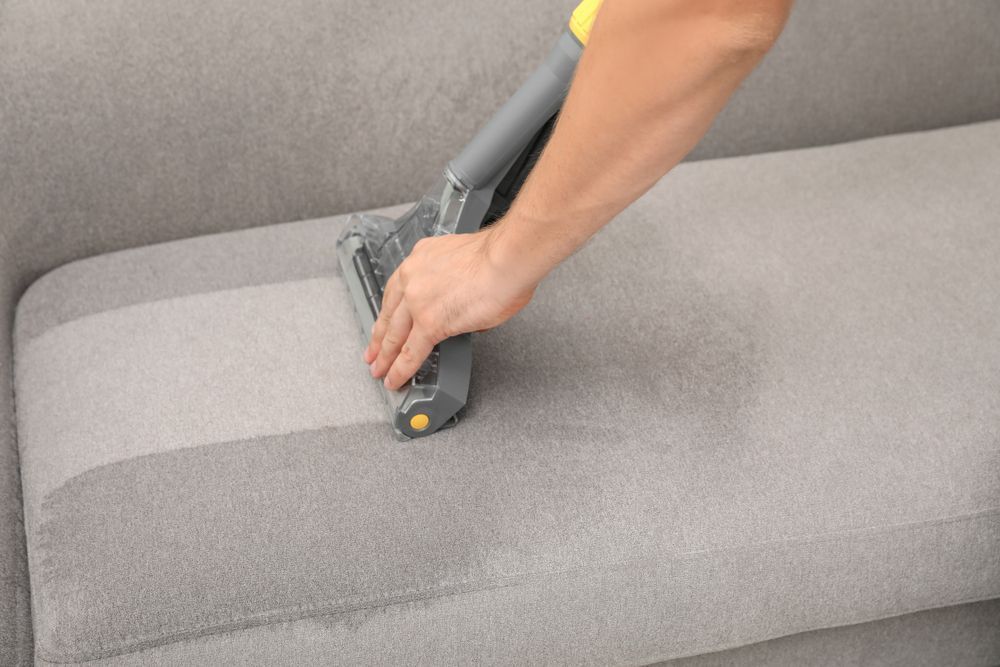 Person Cleaning a Gray Upholstered Sofa With a Handheld — Clean Coast Carpet Cleaning in Port Stephens, NSW
