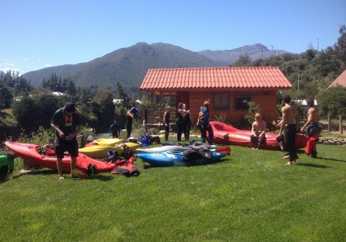 Gente con kayaks en un campo de hierba cerca de una cabaña de techo rojo, con montañas al fondo.