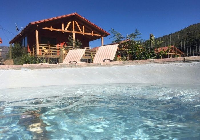 Cabaña frente a la playa, junto a una piscina, con dos tumbonas y agua azul en primer plano.