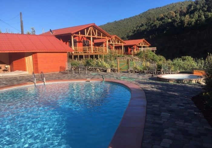 Piscina del complejo con agua azul junto a cabañas de madera con techos rojos y una ladera de fondo al atardecer.