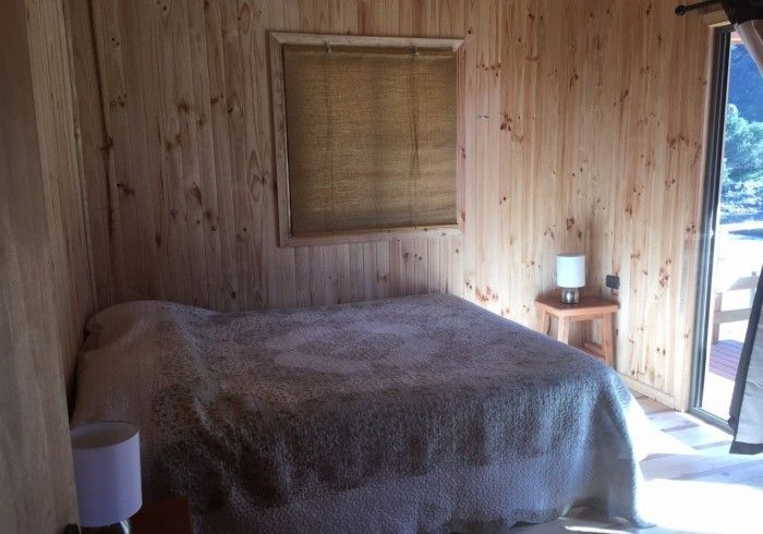 Dormitorio con paneles de madera, cama, mesita de noche pequeña, persiana y puerta abierta con vistas soleadas al exterior.