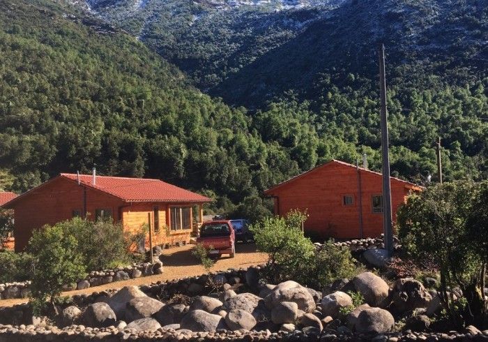Dos cabañas naranjas con un camión rojo, situadas entre rocas y montañas verdes bajo un cielo despejado.