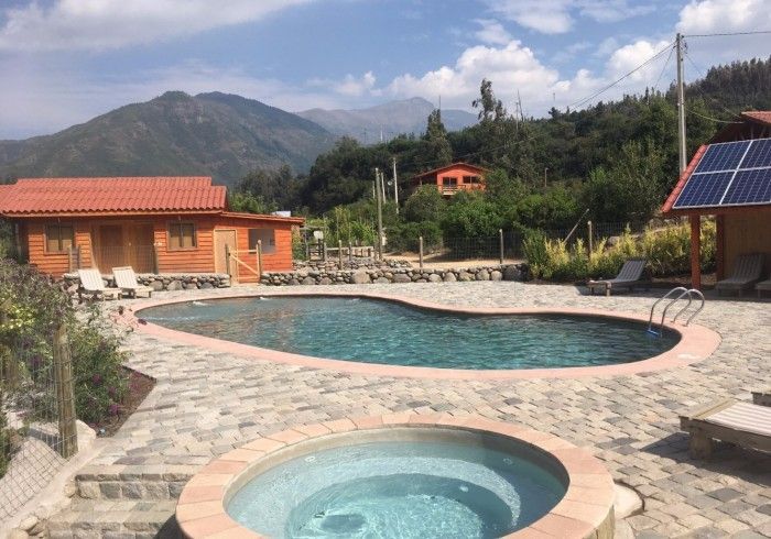 Piscina en un complejo turístico de montaña con jacuzzi, patio de piedra y paneles solares, rodeada de árboles y colinas.