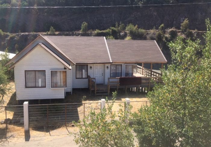 Pequeña casa blanca con porche en la ladera de una colina, rodeada de matorrales secos y árboles.