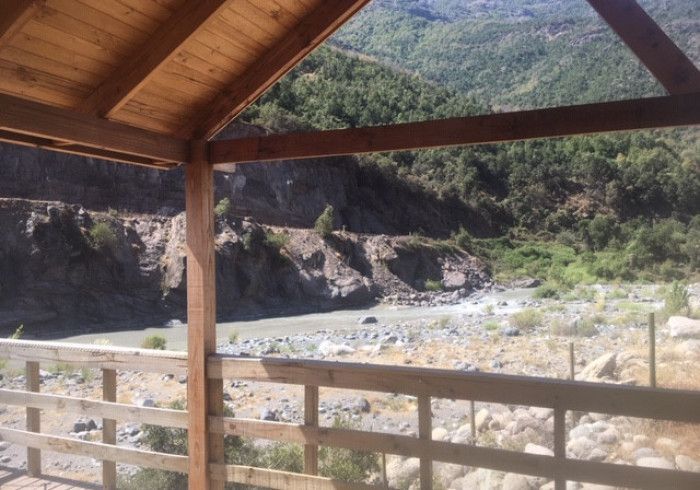 Refugio de madera con vistas a un lecho de río rocoso y una ladera verde bajo la luz del día.
