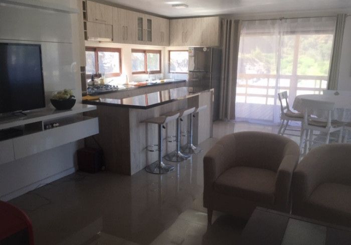 Cocina y sala de estar luminosas con taburetes de bar, sillones y luz natural que entra por los grandes ventanales.