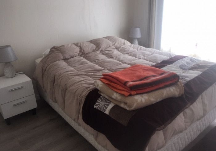 Una cama bien hecha con ropa de cama beige, una manta roja doblada y dos mesitas de noche en un dormitorio luminoso.