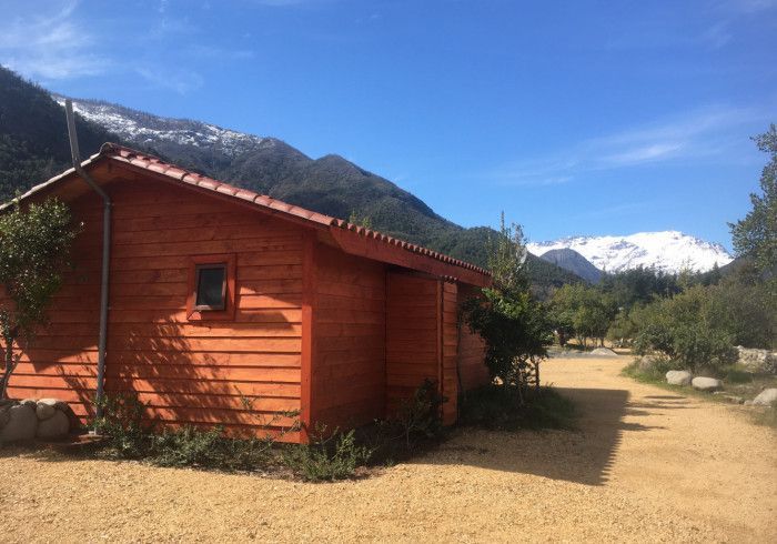 Cabaña roja en un sendero rural soleado con montañas y nieve al fondo.
