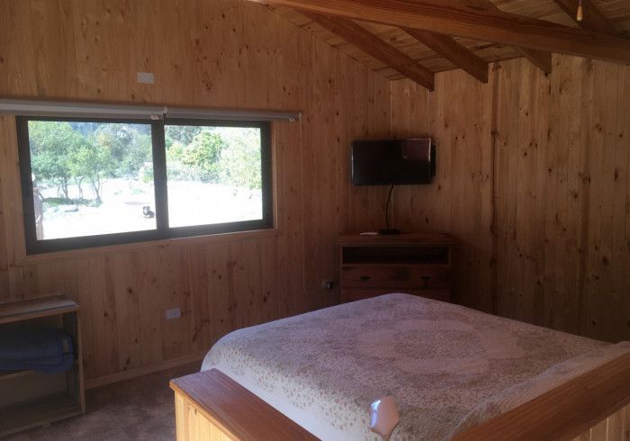 Dormitorio con paneles de madera, cama, ventana, cómoda y televisor montado en la pared.