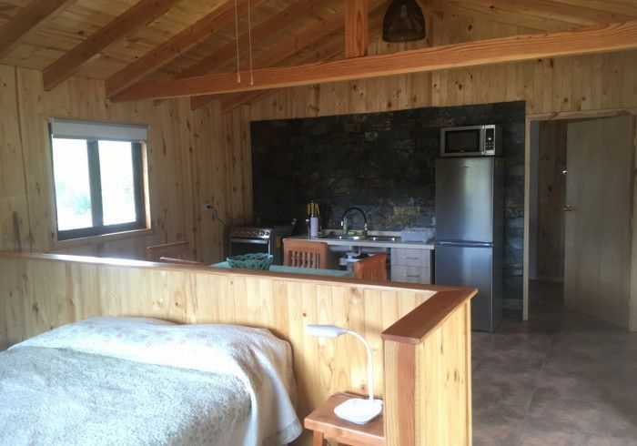 Habitación tipo cabaña con cocina americana, paredes de madera, cama, ventana y refrigerador de acero inoxidable.