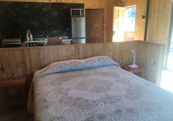 Acogedor dormitorio con una cama en primer plano, paredes revestidas de madera y una zona de cocina al fondo.