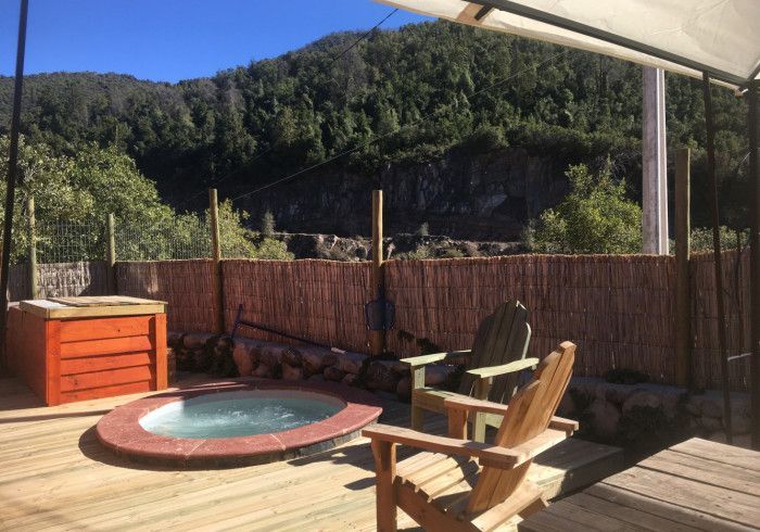 Terraza de madera con jacuzzi, sillas y vistas a la ladera vallada bajo un cielo soleado.