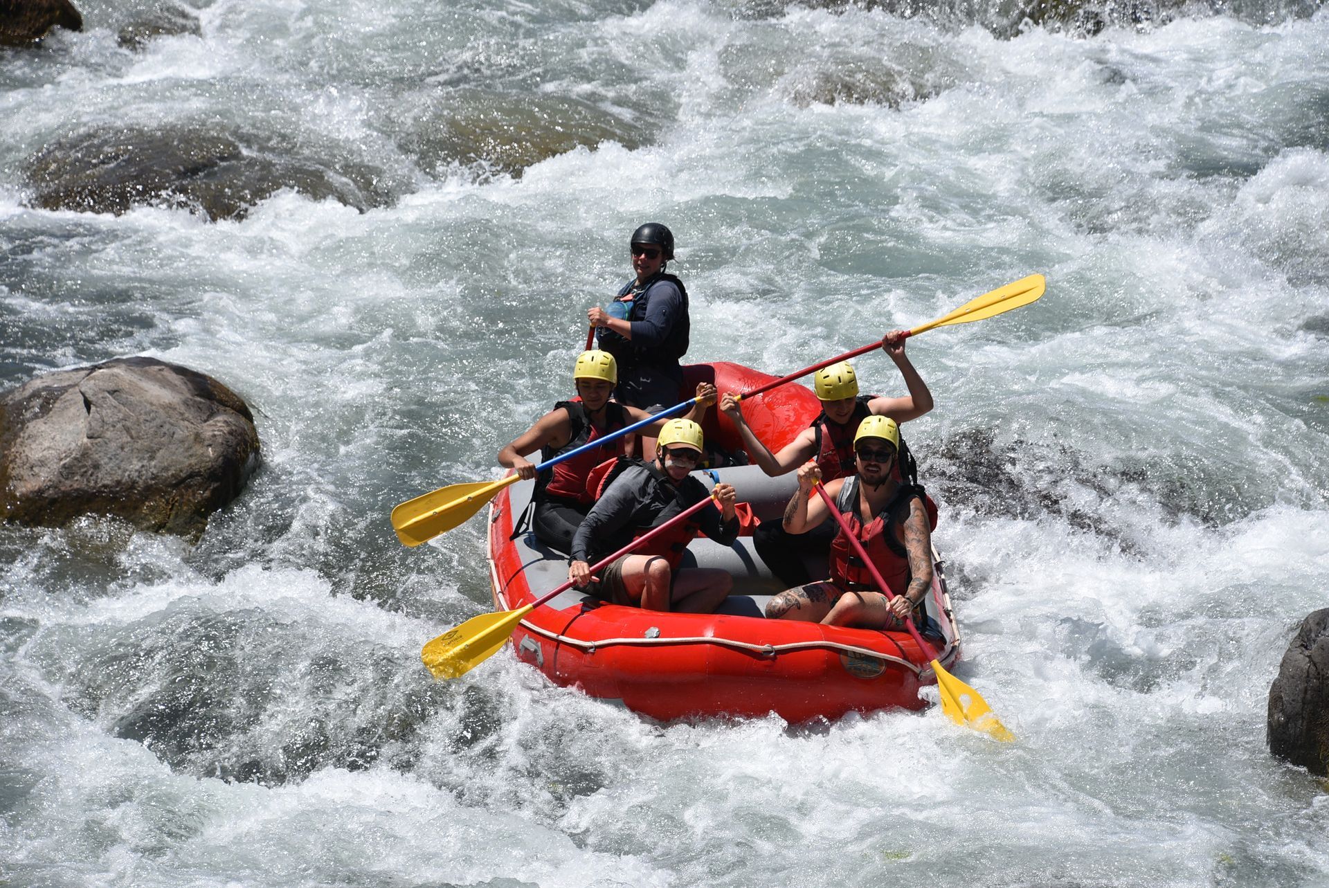 Personas practicando rafting en aguas bravas en una balsa roja a través de rápidos turbulentos, con los remos en alto en el agua salpicada.