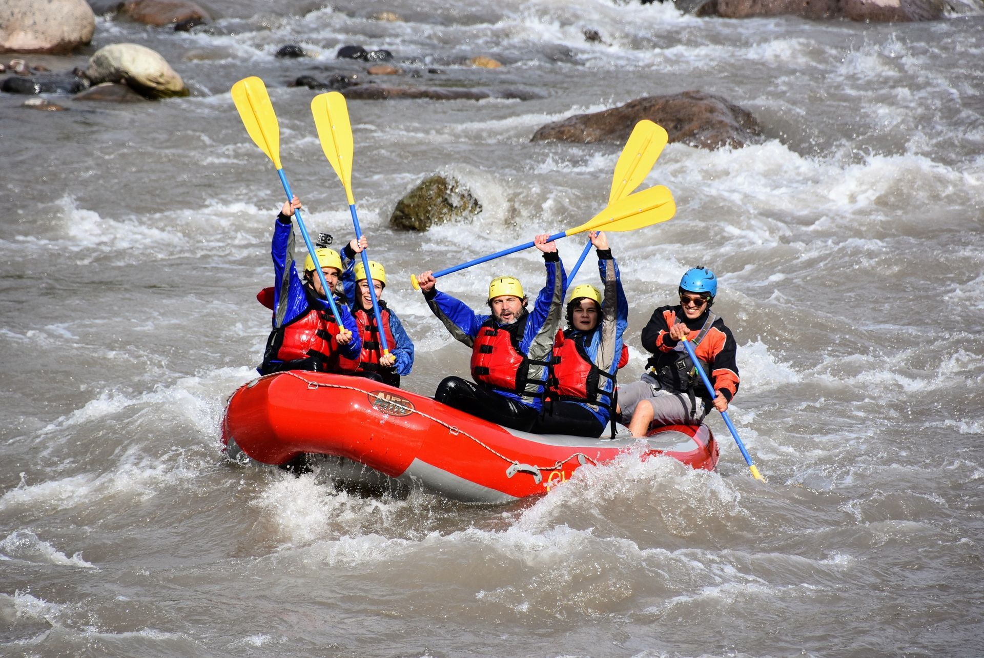 Cuatro personas practicando rafting en aguas bravas en una balsa roja, remando a través de rápidos turbulentos del río.