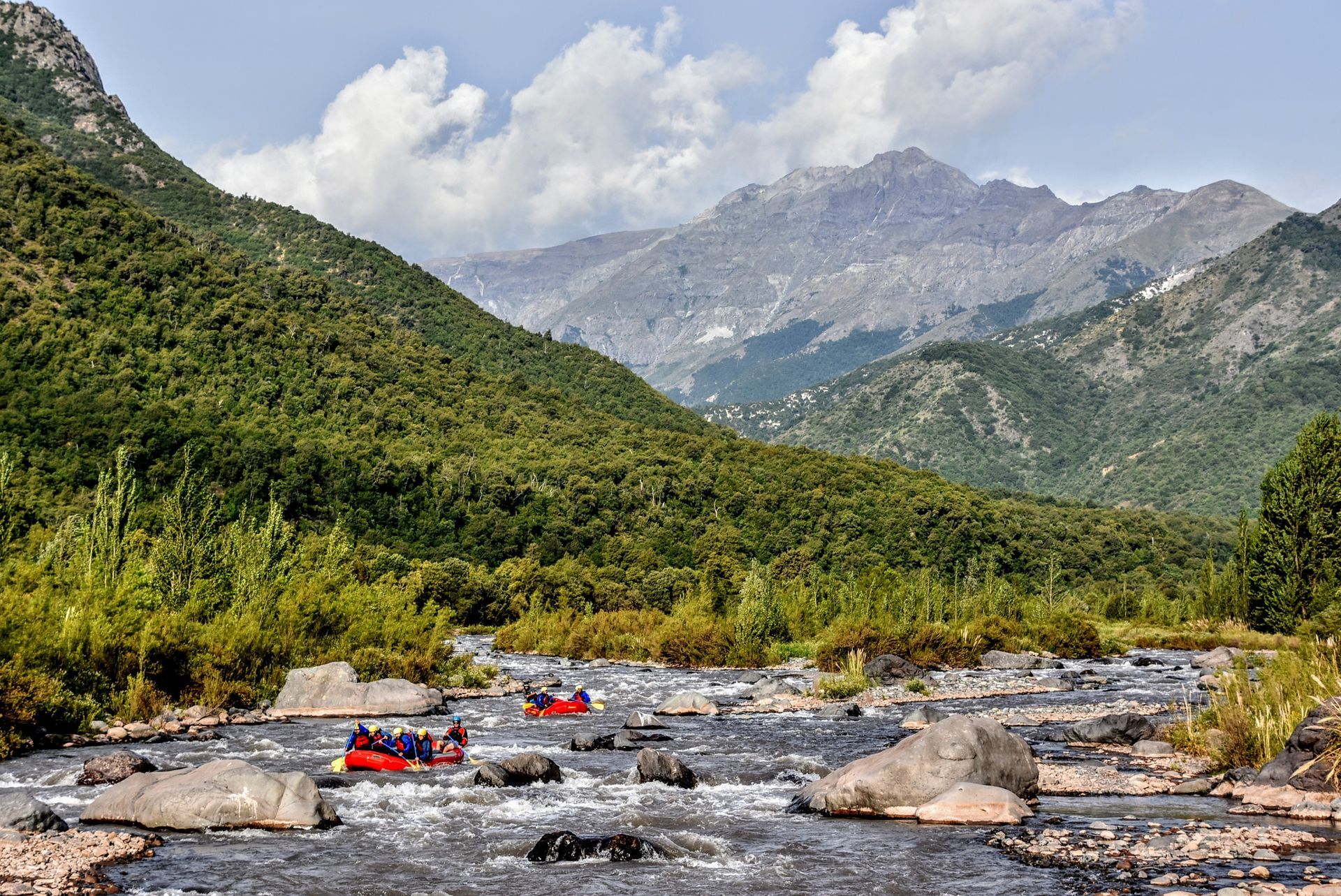 Balseros en balsas rojas en un río rocoso bajo montañas verdes y picos nublados.