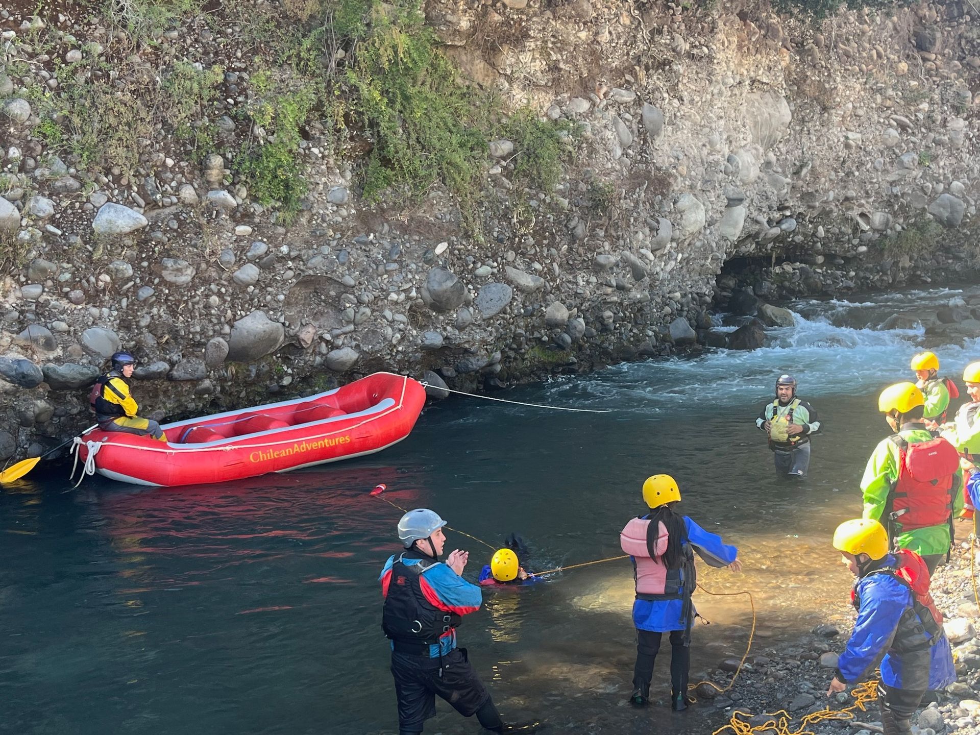 Varias personas con casco permanecen de pie en aguas poco profundas junto a una balsa inflable roja en la orilla rocosa de un río.