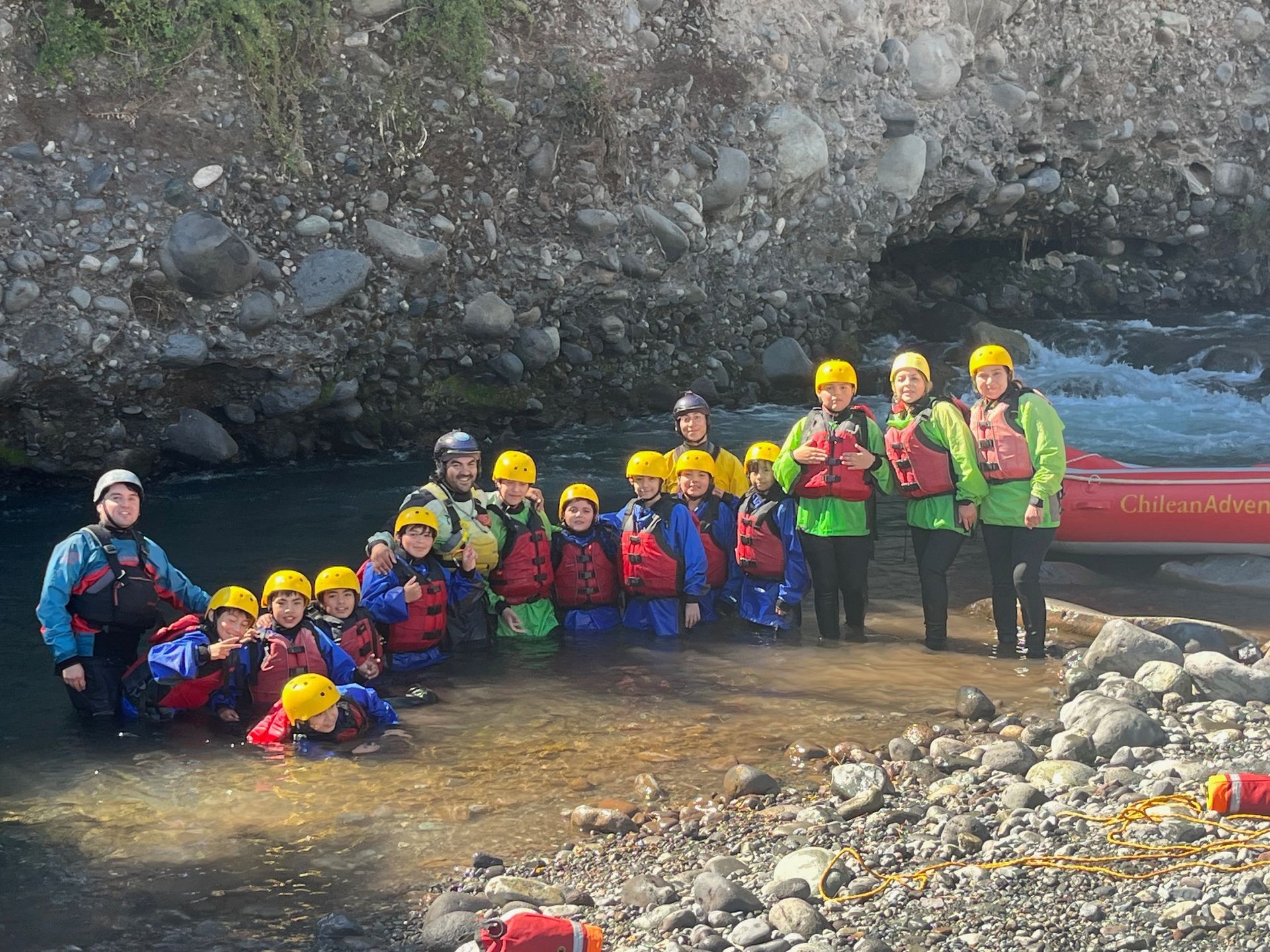 Grupo de personas con cascos y chalecos salvavidas de pie en aguas poco profundas del río, junto a un acantilado rocoso y una balsa.