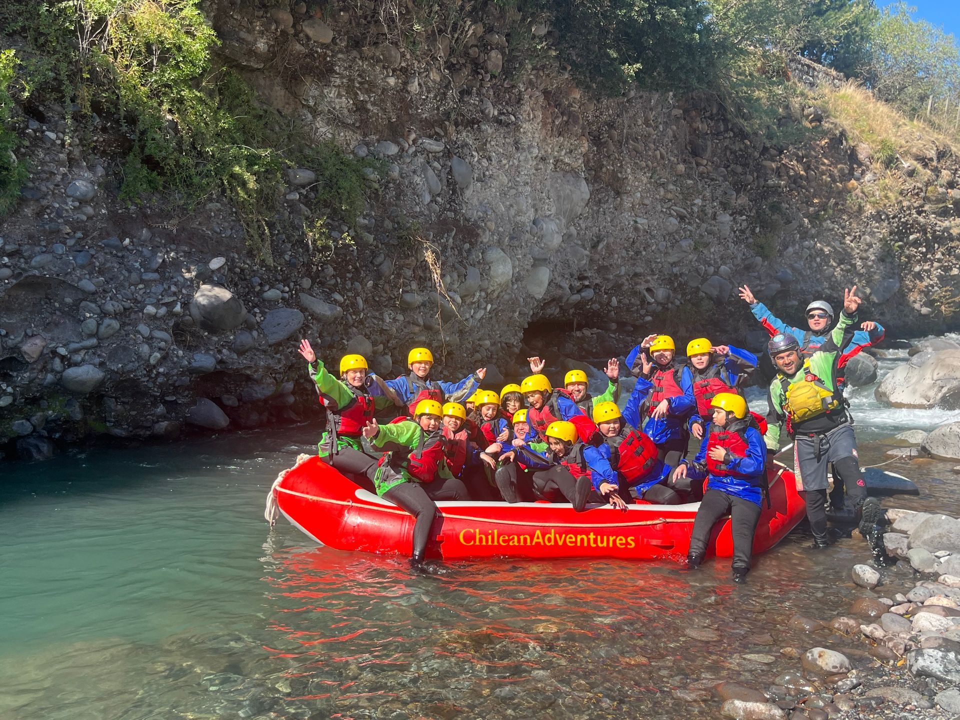 Grupo en una balsa roja con cascos coloridos en un río turquesa junto a un cañón rocoso, una persona saluda
