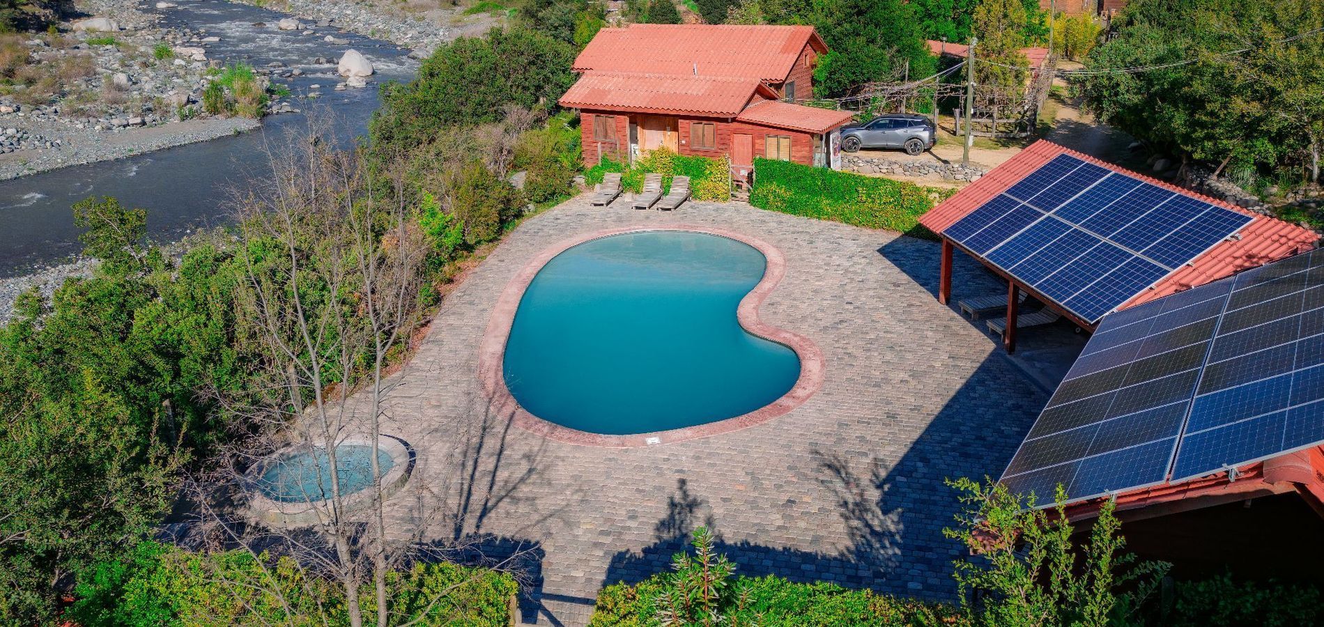 Vista aérea de una piscina con forma de riñón junto a una cabaña de techo rojo, paneles solares y una ribera rocosa.