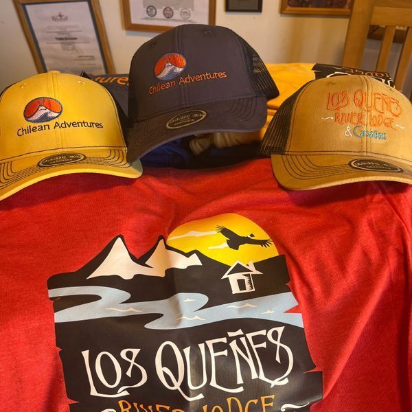 Tres gorras de Los Queñes Adventure sobre una sudadera roja de Los Queñes, expuestas sobre una mesa.