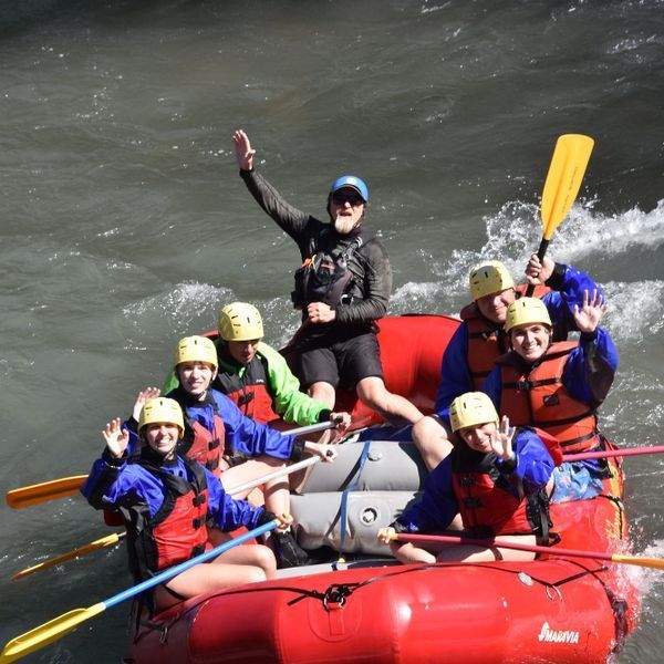 Grupo de personas en una balsa roja navegando por aguas turbulentas, saludando y remando a través de los rápidos.