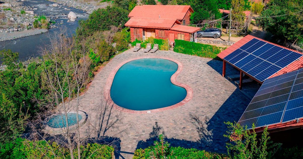 Vista aérea de una piscina en el patio trasero junto a una pequeña casa de techo rojo y paneles solares en un entorno rural.