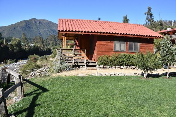 Cabaña roja con techo de tejas rojas, césped verde y paisaje montañoso de fondo bajo un cielo despejado.