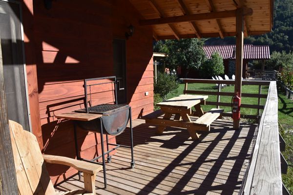 Porche de madera con sillas y mesa, bañado por la luz del sol, con vistas a un jardín con césped y árboles.