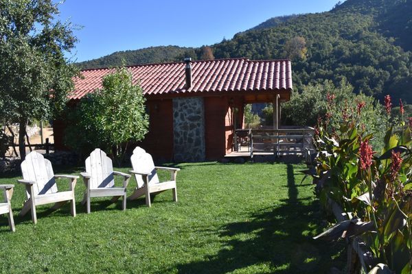 Jardín soleado con sillas blancas sobre césped verde junto a una pequeña casa de piedra y colinas boscosas.