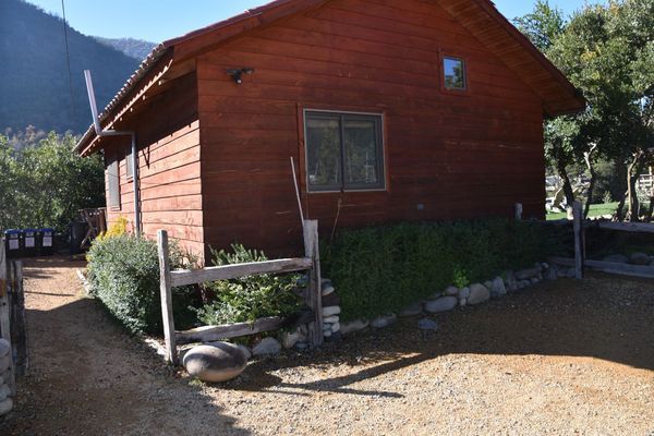 Cabaña de madera roja con un pequeño patio cercado y un camino de grava en un entorno rural montañoso.
