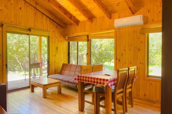 Sala de estar de cabaña con paneles de madera, sofá, mesa de comedor y grandes ventanales con vistas a los árboles.