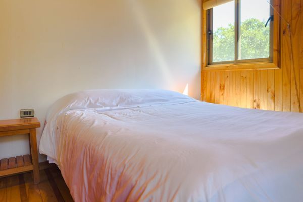 Habitación pequeña con cama blanca, paredes de madera, mesita de noche y ventana con luz natural.
