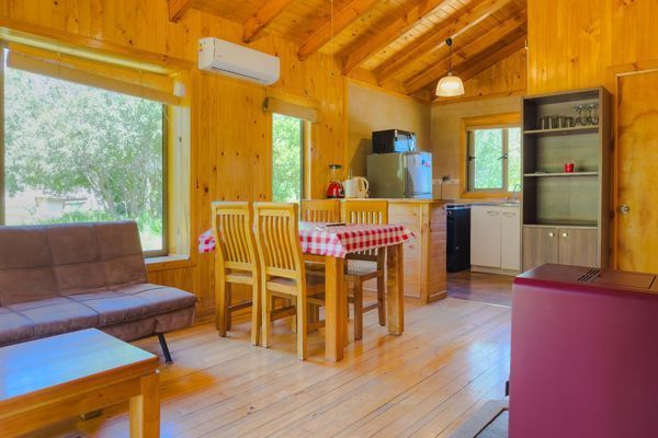 Interior de cabaña de madera con mesa de comedor, sofá, cocina y grandes ventanales con vistas a la vegetación.