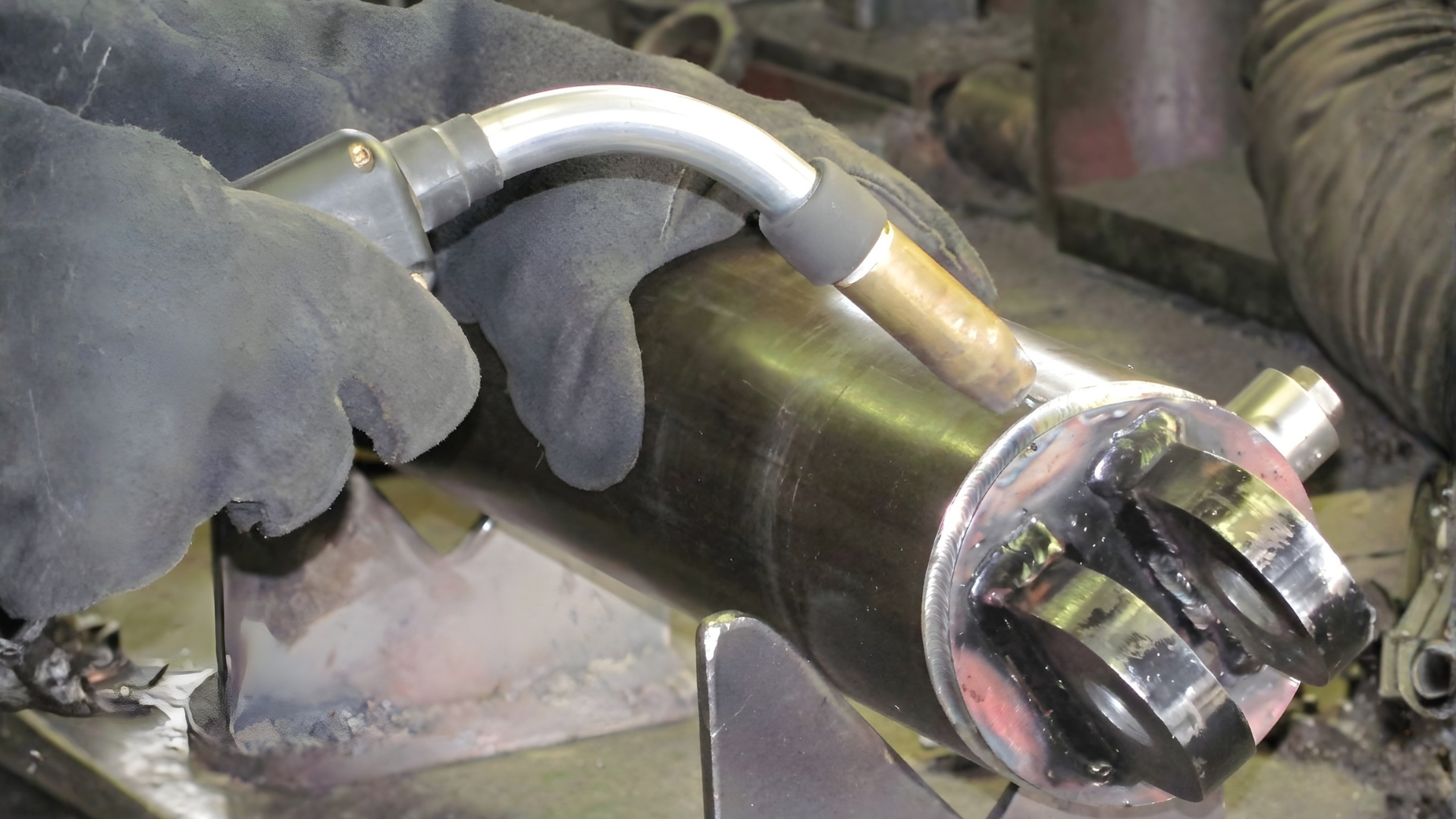 Gloved hands use a torch to cut a metal pipe, with sparks and a vise holding it in place.