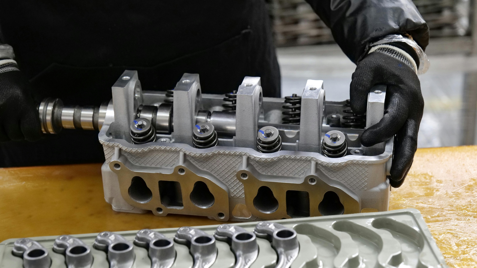 Mechanic holding a metal engine cylinder head on a workbench, with another cylinder head below.