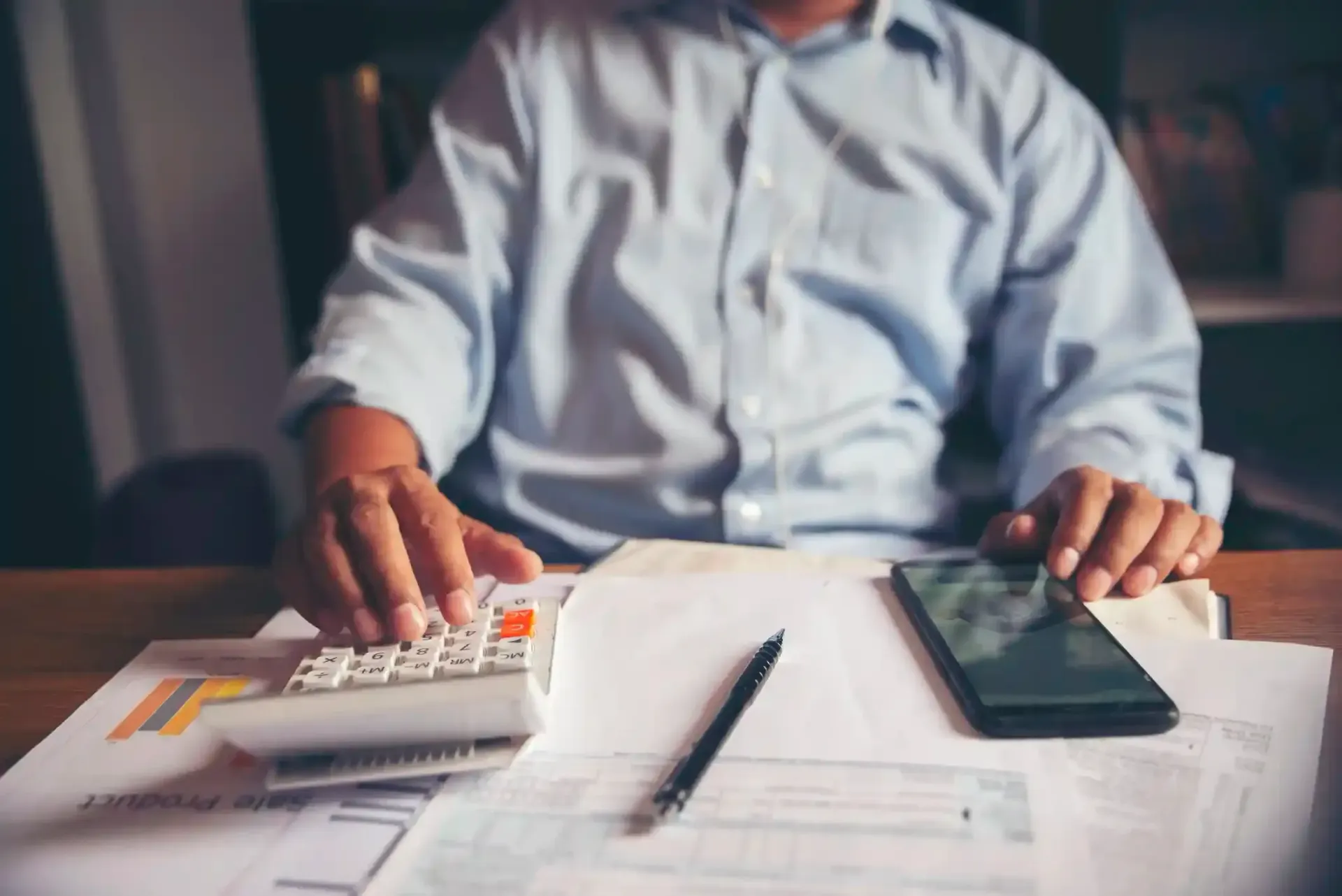 Person using a calculator and smartphone on a desk with paperwork; focused on finances.