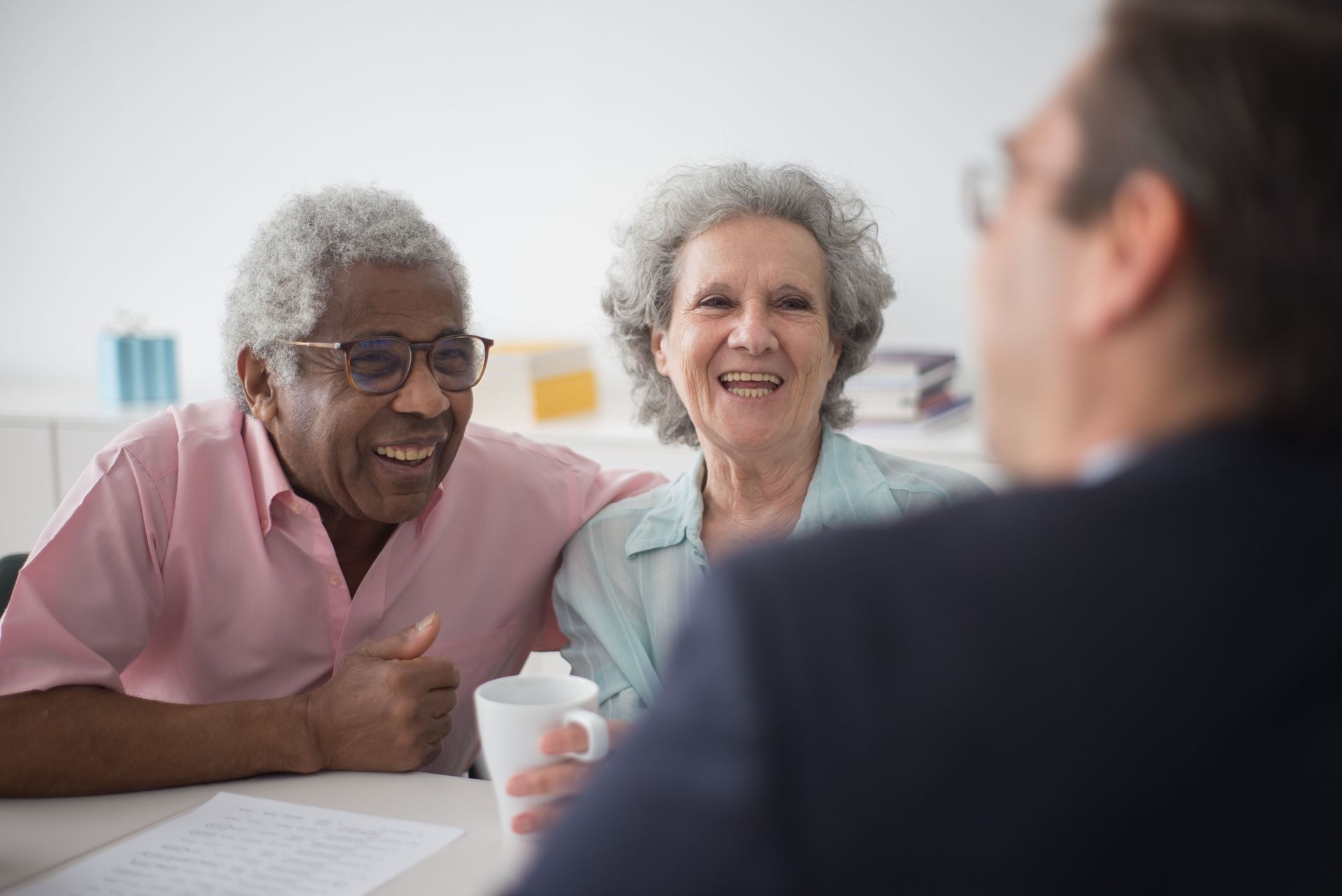 Two smiling people at a table speaking with a person in the foreground, in a bright room.
