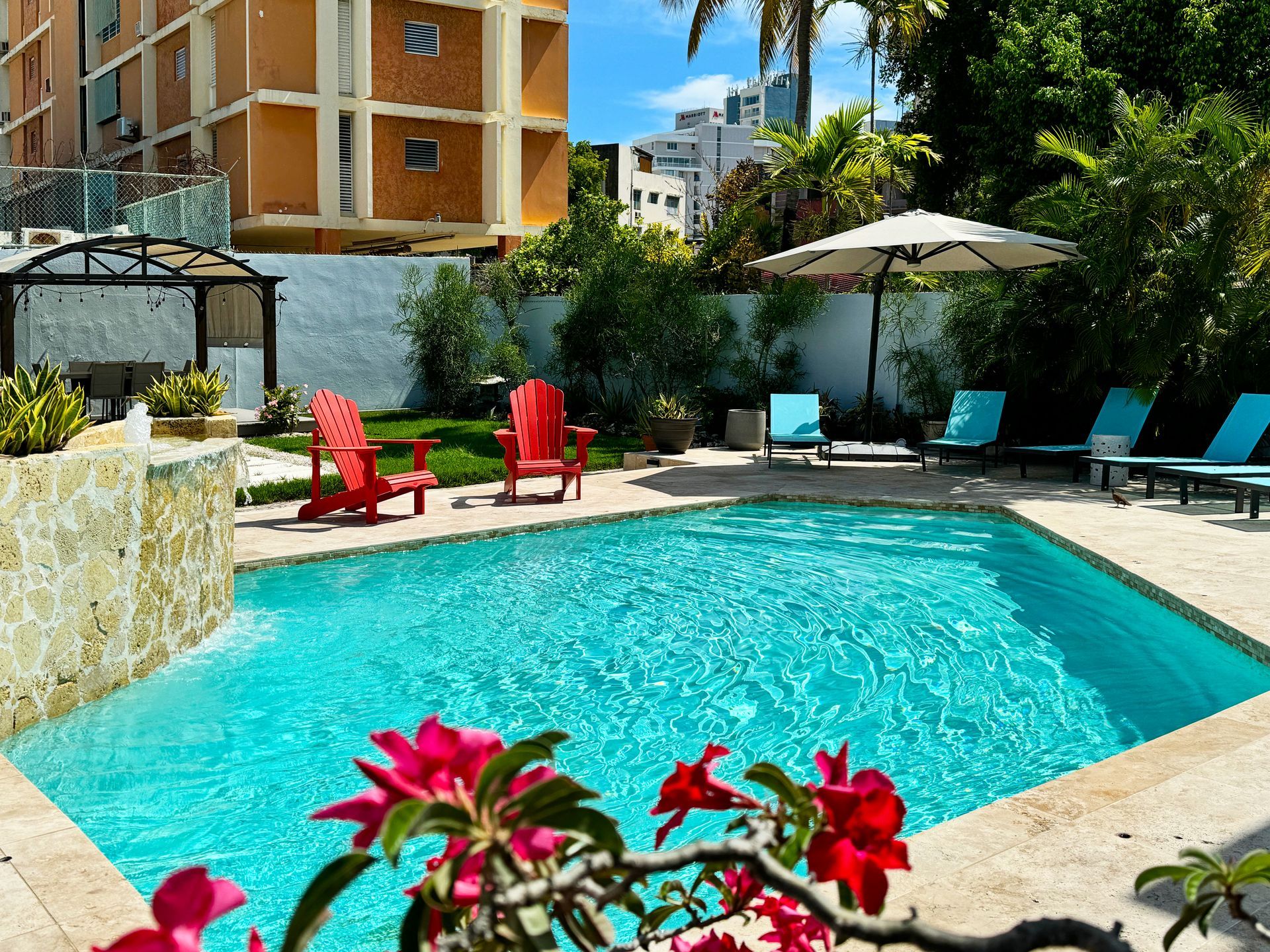 Sunny poolside courtyard with turquoise water, red chairs, blue loungers, and flowering plants