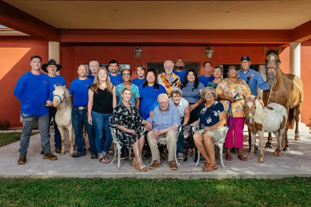 Group portrait of people and horses posing on a patio. Many wear blue, while a man and woman are seated in the foreground.