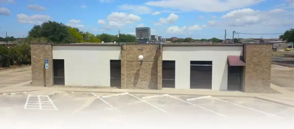 A low, light-colored building with stone accents and dark doors sits under a blue sky. It has a small parking area.