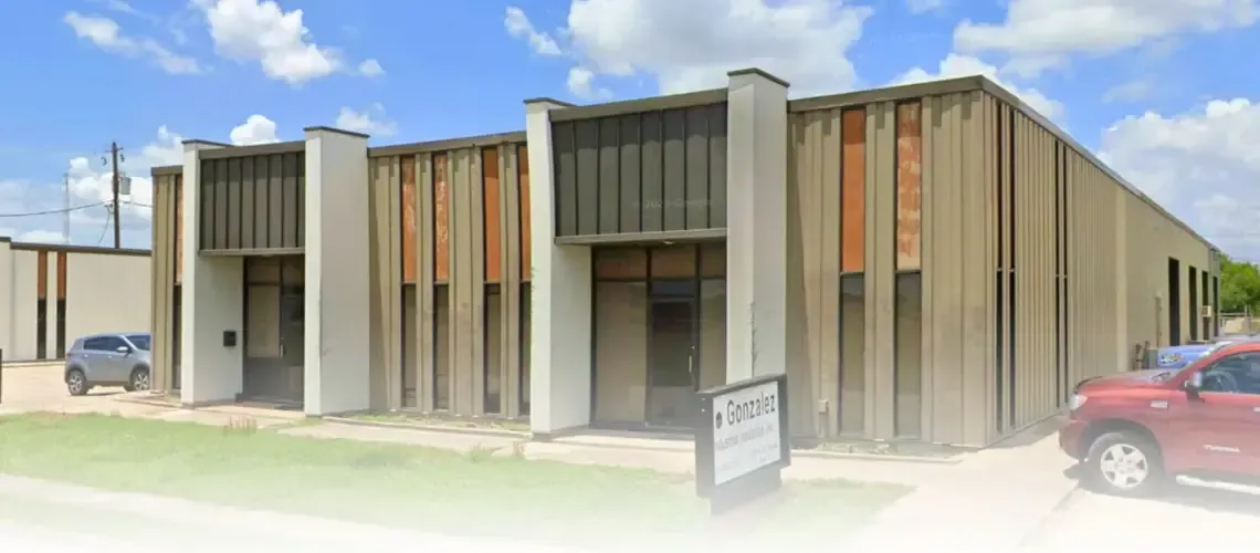 A two-story beige commercial building with multiple entrances, a sign in front, and a blue sky background. Two cars are parked nearby.