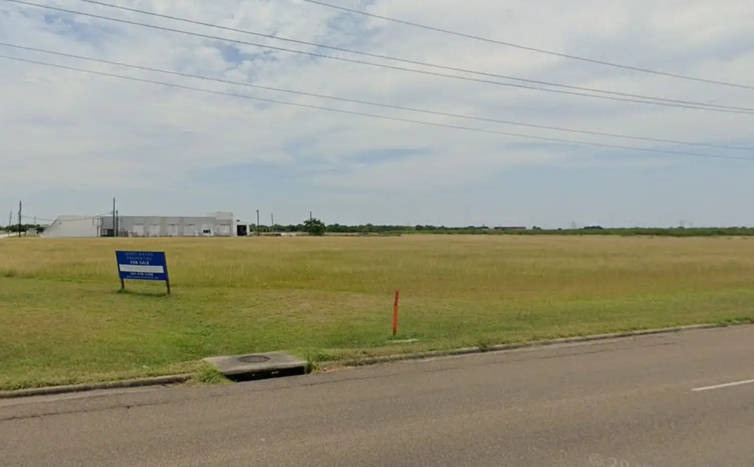Grassy field with a building in the background, sign, and a sewer grate on the roadside. Cloudy sky.