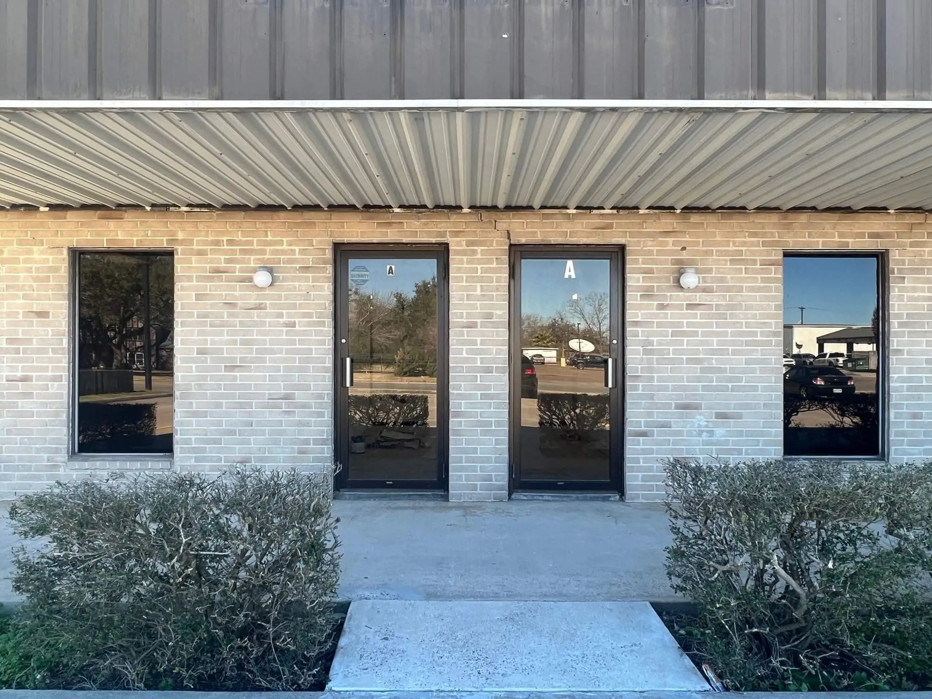 Exterior view of a building with two doors and two windows, all with dark frames, and a brick facade. Bushes are at the bottom.