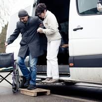 Man in a wheelchair being helped onto a vehicle by another person.